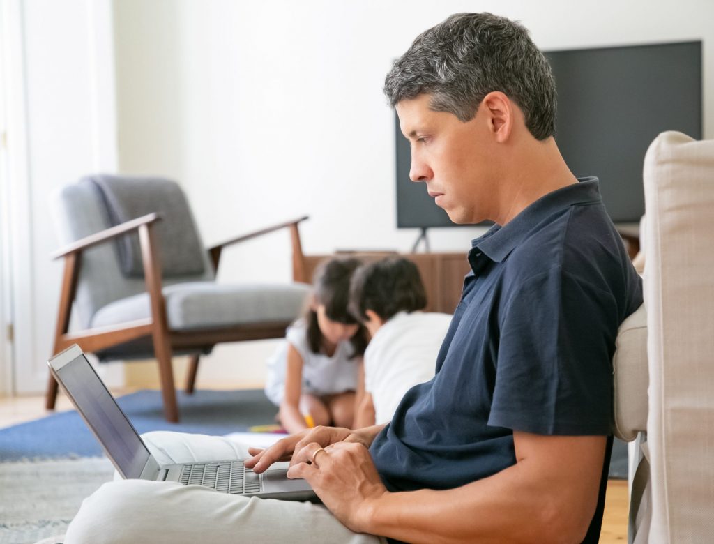 Serious professional working at home, sitting on floor and using laptop while his little kids playing in background. Distance work or home office concept